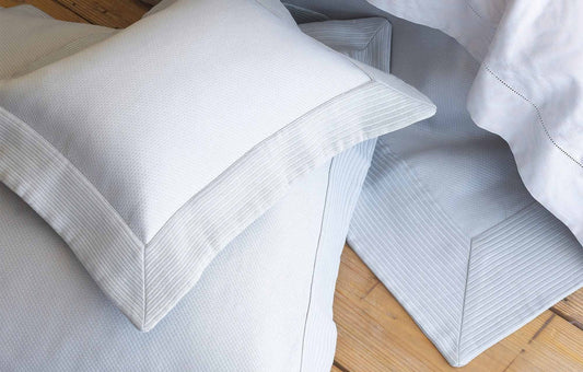 Light gray textured pillow sham with a wide, ribbed border resting on a wooden floor next to a bed.