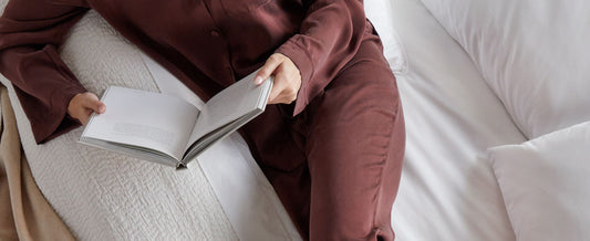Woman in burgundy lounge wear on a white bed, reading a book
