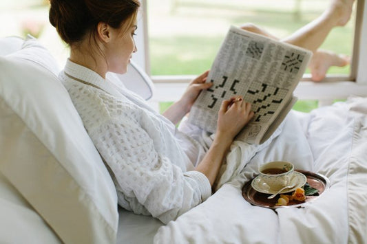 A woman in a robe does the newspaper crossword in bed with a cup of tea beside her