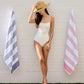 Woman in a white swimsuit standing between two Aegean Beach Towels against a brick wall, Navy, Coral