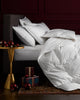 A white duvet cover and matching pillow shams arranged on a bed in a dark bedroom setting, with a gold side table, red ornaments and wrapped gifts.