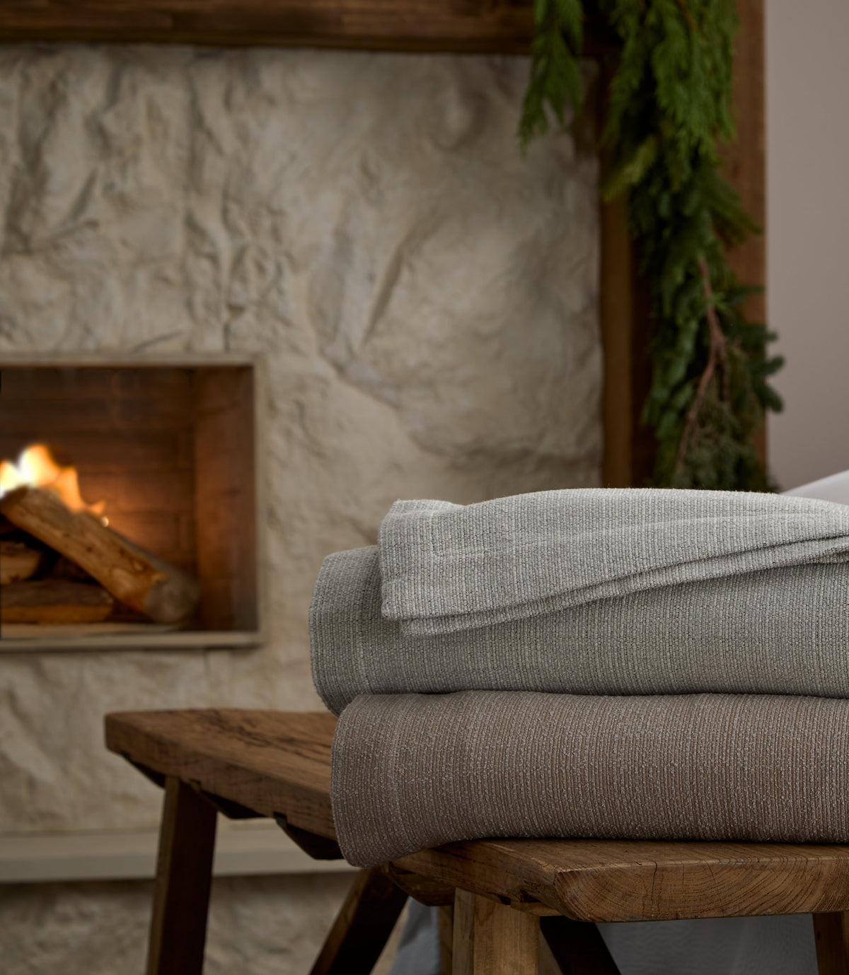 Hampton Throw Blankets stacked on top a wooden stool next to a fireplace, Gray, Mushroom