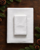 A neatly folded white bedding set, including a fitted sheet and a pillowcase, surrounded by pinecones.