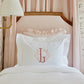 Soprano Sateen Sham in White featuring a monogram, displayed on a bed with a light pink striped headboard.