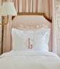 Soprano Sateen Sham in White featuring a monogram, displayed on a bed with a light pink striped headboard.