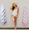 Woman in a white swimsuit with a straw hat standing between two beach towels against a light wooden wall.