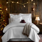 A bed dressed in Hamilton Quilted Coverlet styled in a cozy cabin, with a lit Christmas tree, and a bedside lamp.