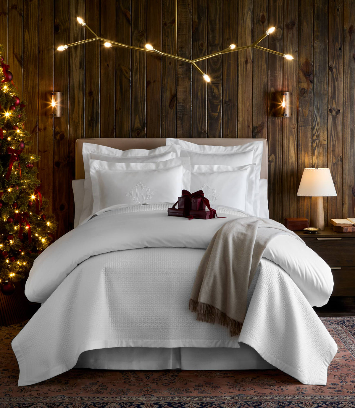 A bed dressed in Hamilton Quilted Coverlet styled in a cozy cabin, with a lit Christmas tree, and a bedside lamp.