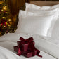 Hamilton Coverlet and Virtuoso Sateen Sham in White monogrammed and styled on a white bed, next to a lit Christmas tree. 