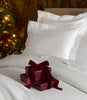 Hamilton Coverlet and Virtuoso Sateen Sham in White monogrammed and styled on a white bed, next to a lit Christmas tree. 
