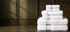 A stack of white cotton towels in various sizes, neatly folded and arranged on a dark wooden counter against a dark olive green paneled wall with soft window light shadows.